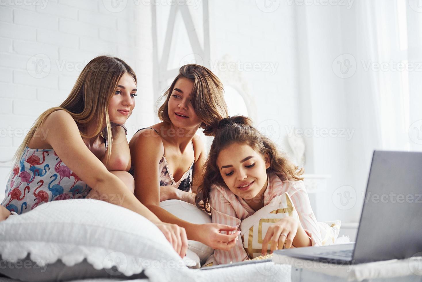 Watching movie on laptop and eating popcorn. Happy female friends having good time at pajama party in the bedroom photo