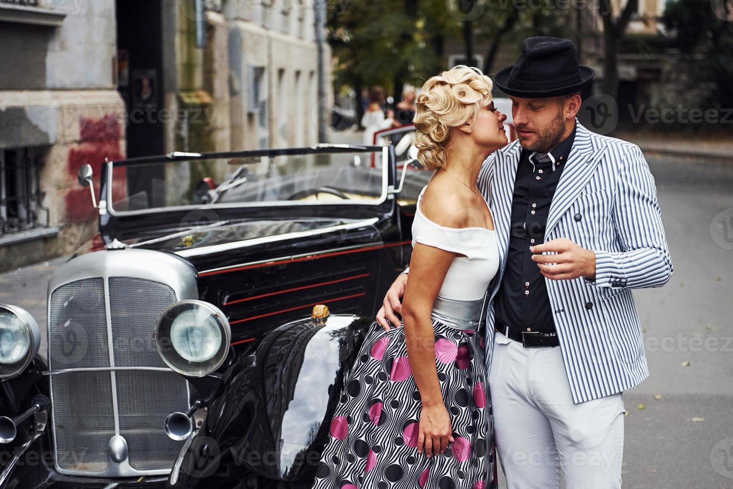 una pareja hermosa con ropa antigua está en la ciudad con un auto retro foto