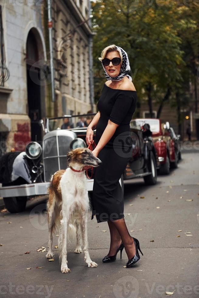 Blonde woman in sunglasses and in black dress near old vintage classic car with her dog photo