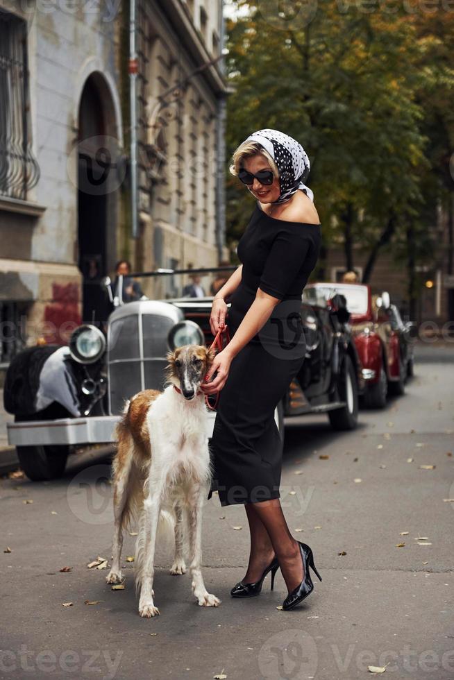 Blonde woman in sunglasses and in black dress near old vintage classic car with her dog photo