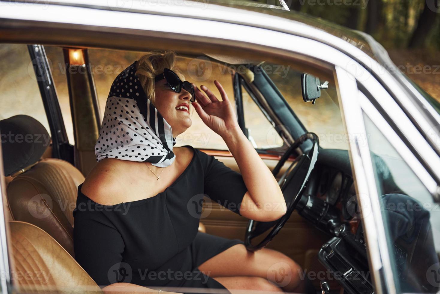 Blonde woman in sunglasses and in black dress sits in old vintage classic car photo