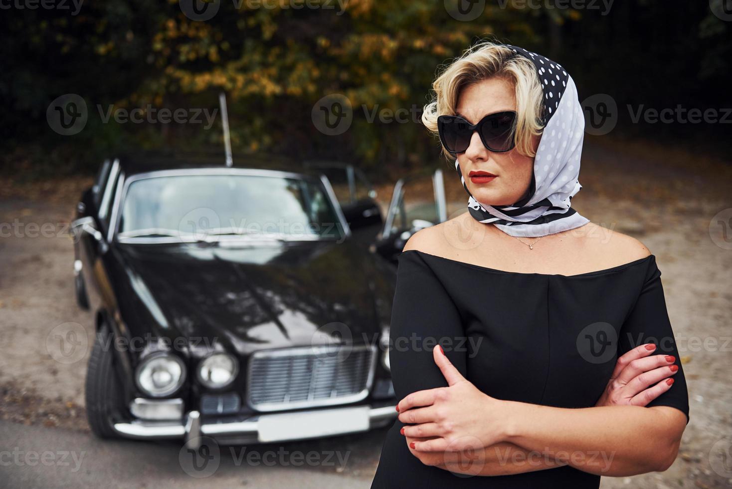 Blonde woman in sunglasses and in black dress near old vintage classic car photo