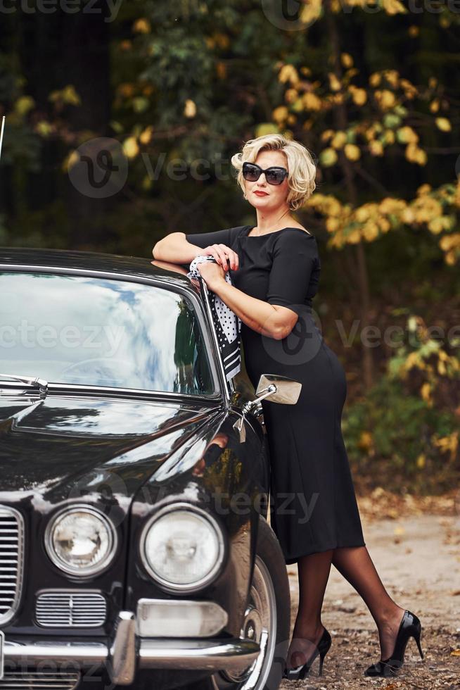 mujer rubia con gafas de sol y vestido negro cerca del viejo coche clásico vintage foto