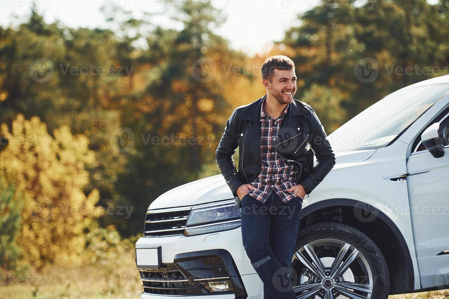 Man in black leather jacket stands near his parked white car outdoors and waits for help photo