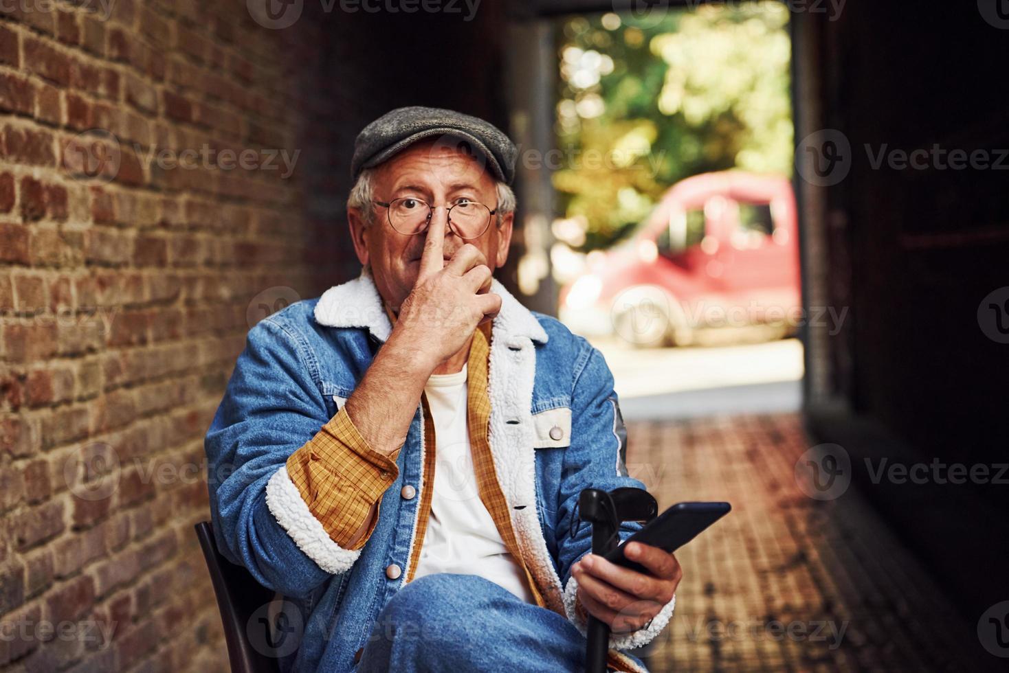 Stylish senior in fashionable clothes and in glasses sits on the chair and uses phone photo