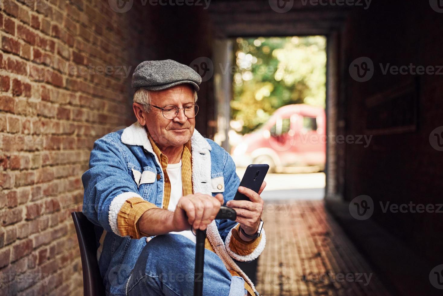 Stylish senior in fashionable clothes and in glasses sits on the chair and uses phone photo
