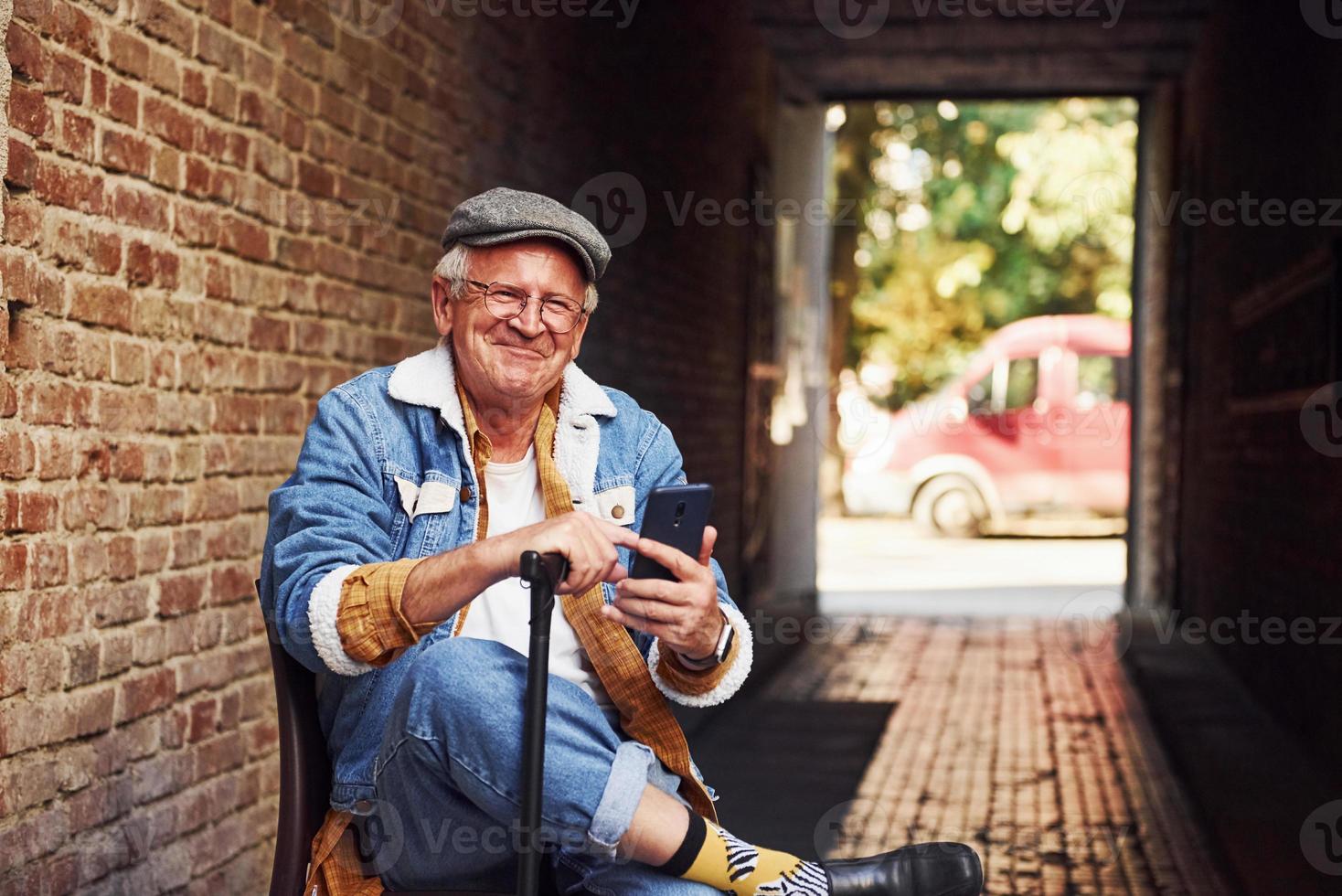 Stylish senior in fashionable clothes and in glasses sits on the chair and uses phone photo