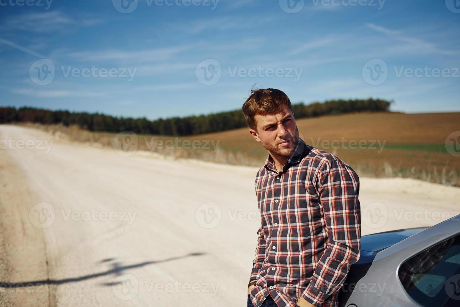 Man in casual clothes leaning on his car outdoors on the road. Rural scene photo