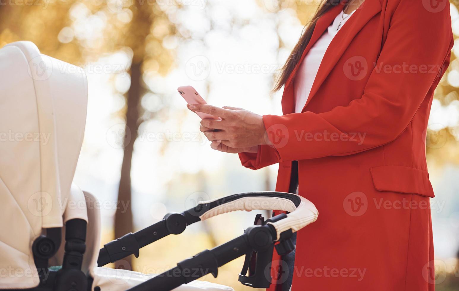 con el teléfono en la mano. madre con abrigo rojo da un paseo con su hijo en el cochecito del parque en otoño foto