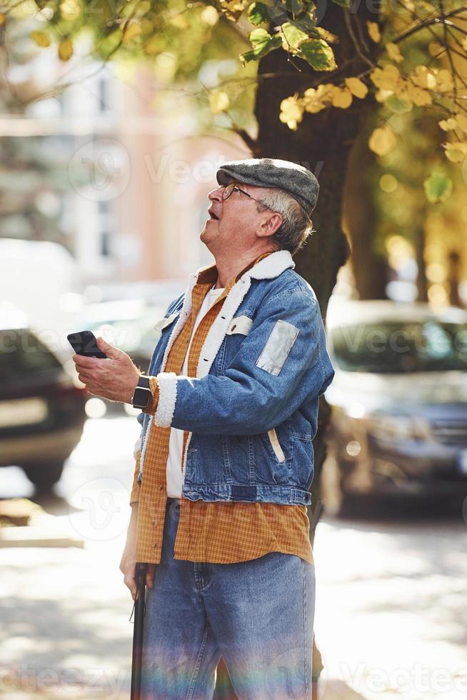 Stylish senior in fashionable clothes and in glasses stands with phone in the city photo