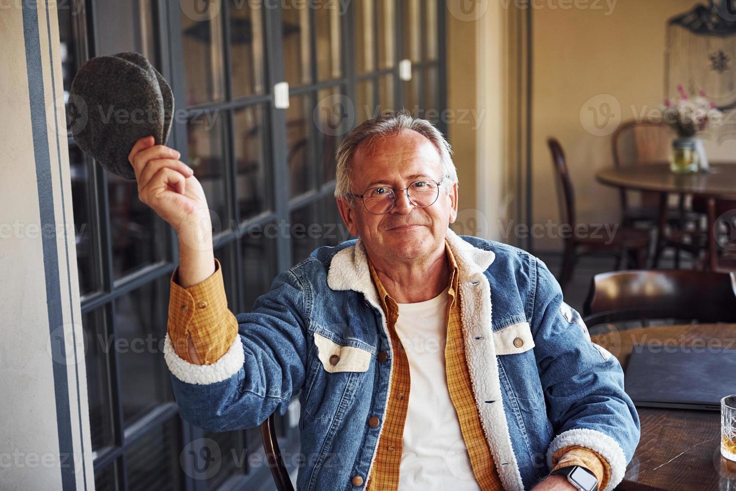 senior con estilo en ropa de moda y con gafas se sienta en el café y sostiene el sombrero foto