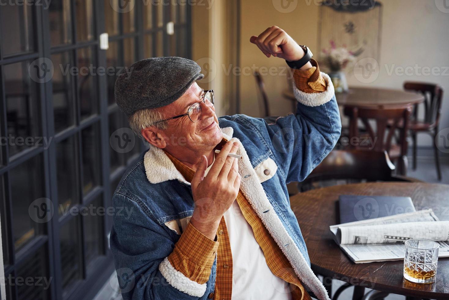 Stylish senior in fashionable clothes and in glasses sits in the cafe with cigarette and talks photo