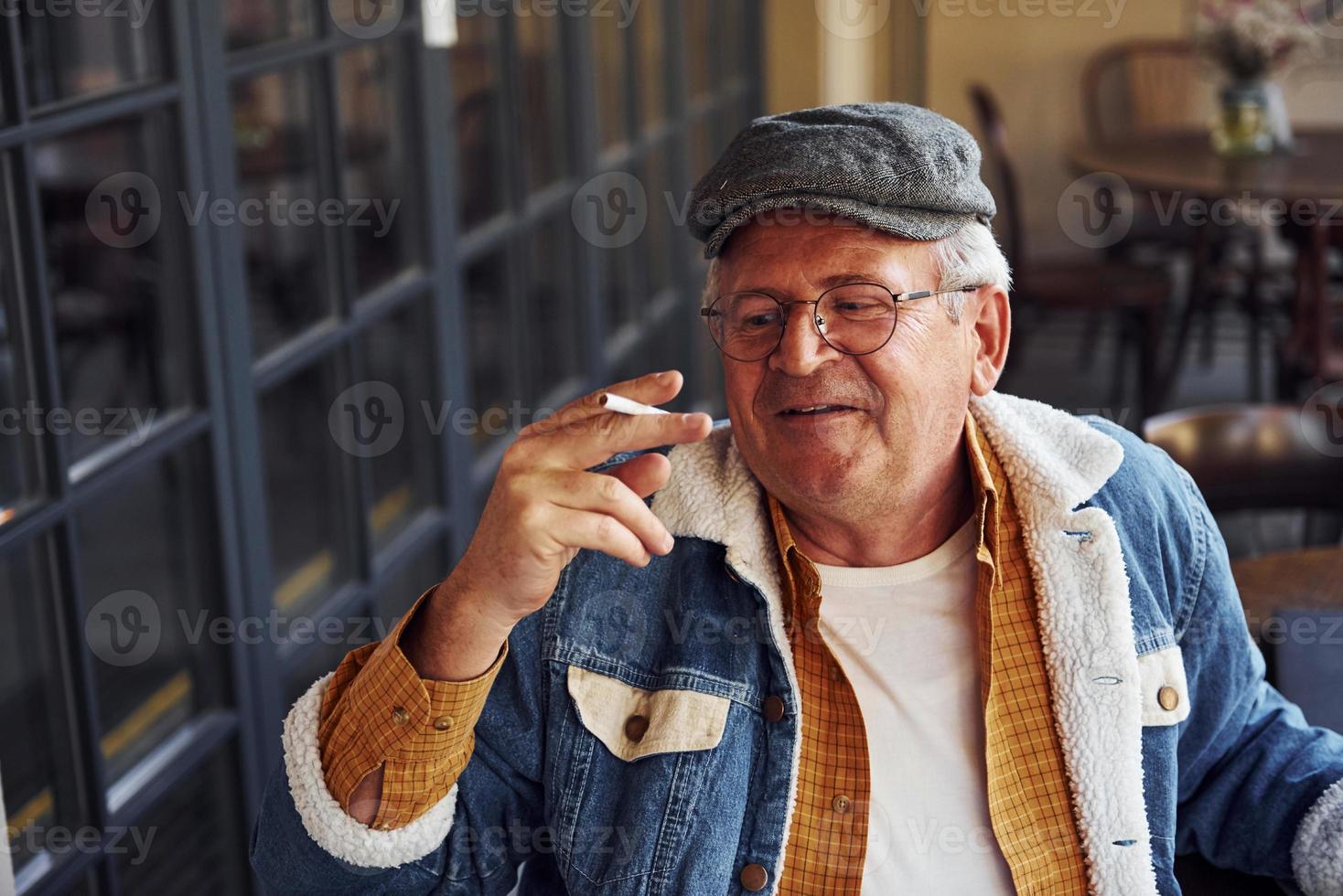 senior con estilo en ropa de moda y con gafas se sienta en el café con cigarrillos y charlas foto
