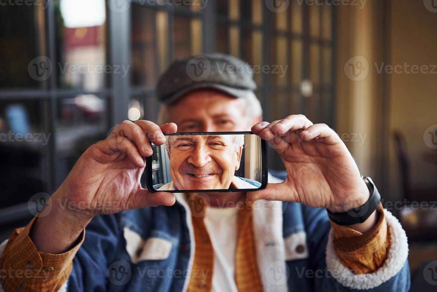 Stylish senior in fashionable clothes and in glasses sits in the cafe and holds phone against his face photo