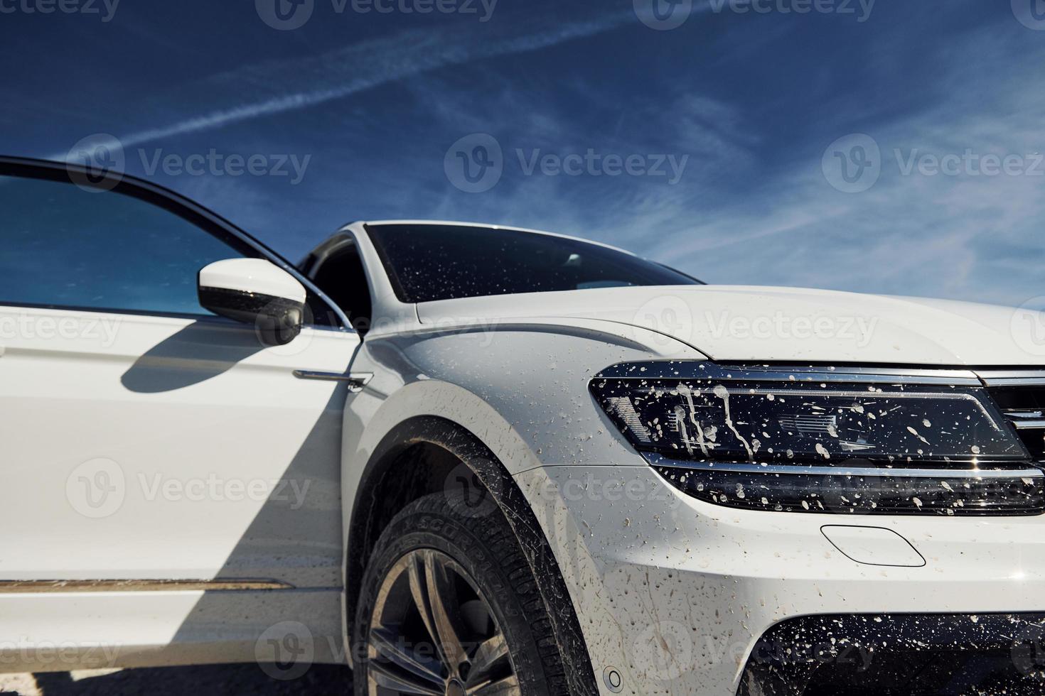 Front view of white modern car parked outdoors against blue sky at background and with dirt on headlights photo
