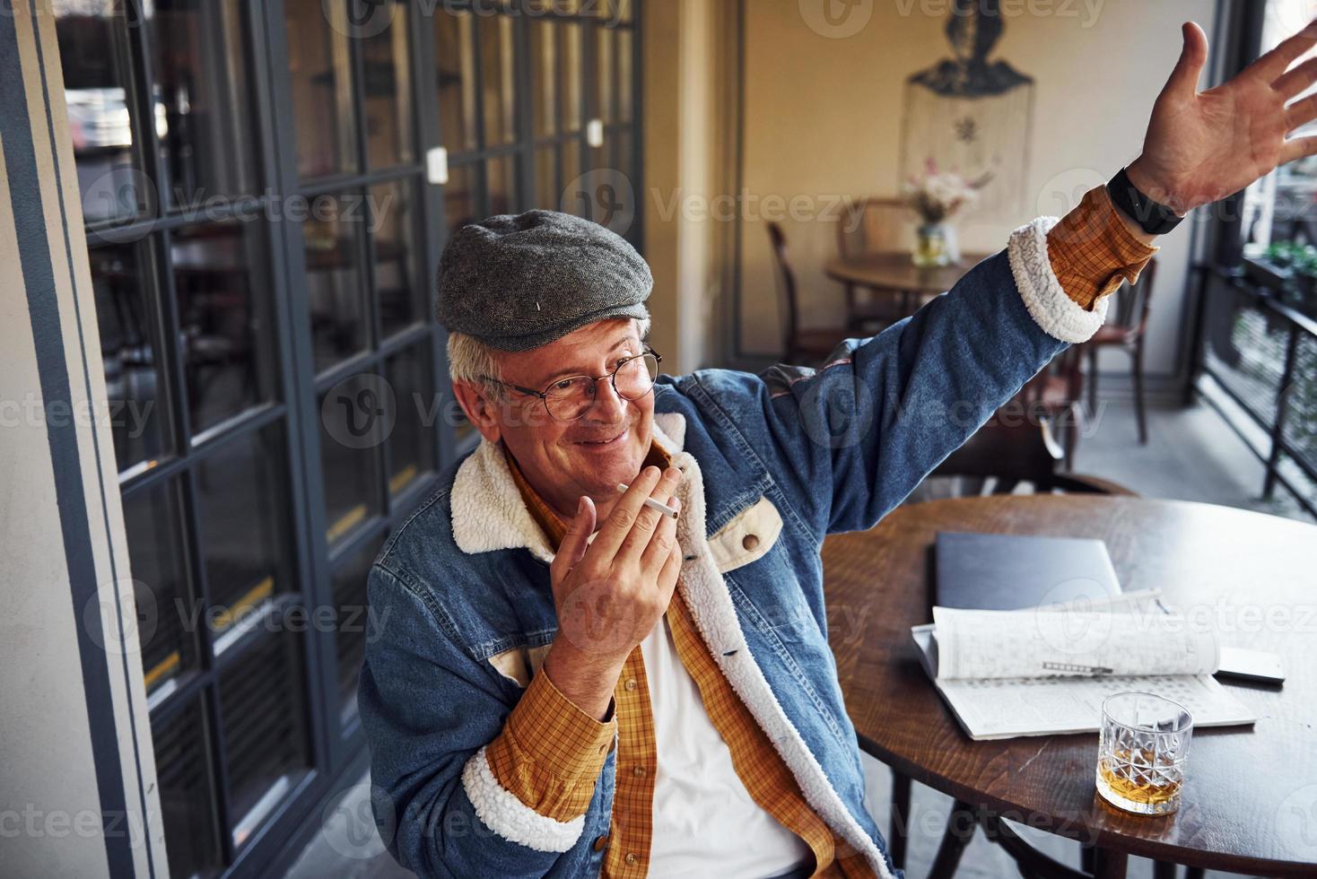 Stylish senior in fashionable clothes and in glasses sits in the cafe with cigarette and talks photo