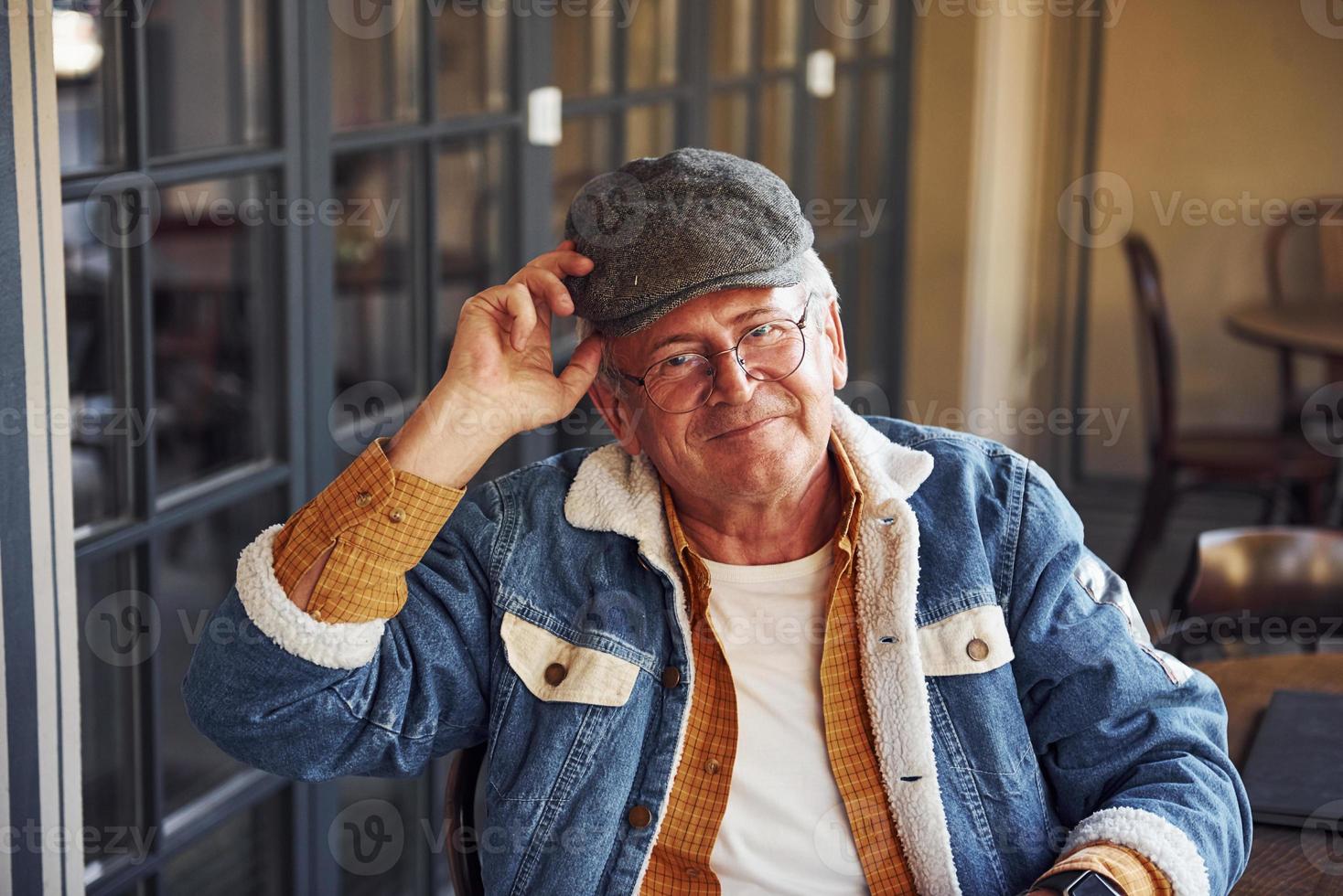 Stylish senior in fashionable clothes and in glasses sits in the cafe and holds hat photo