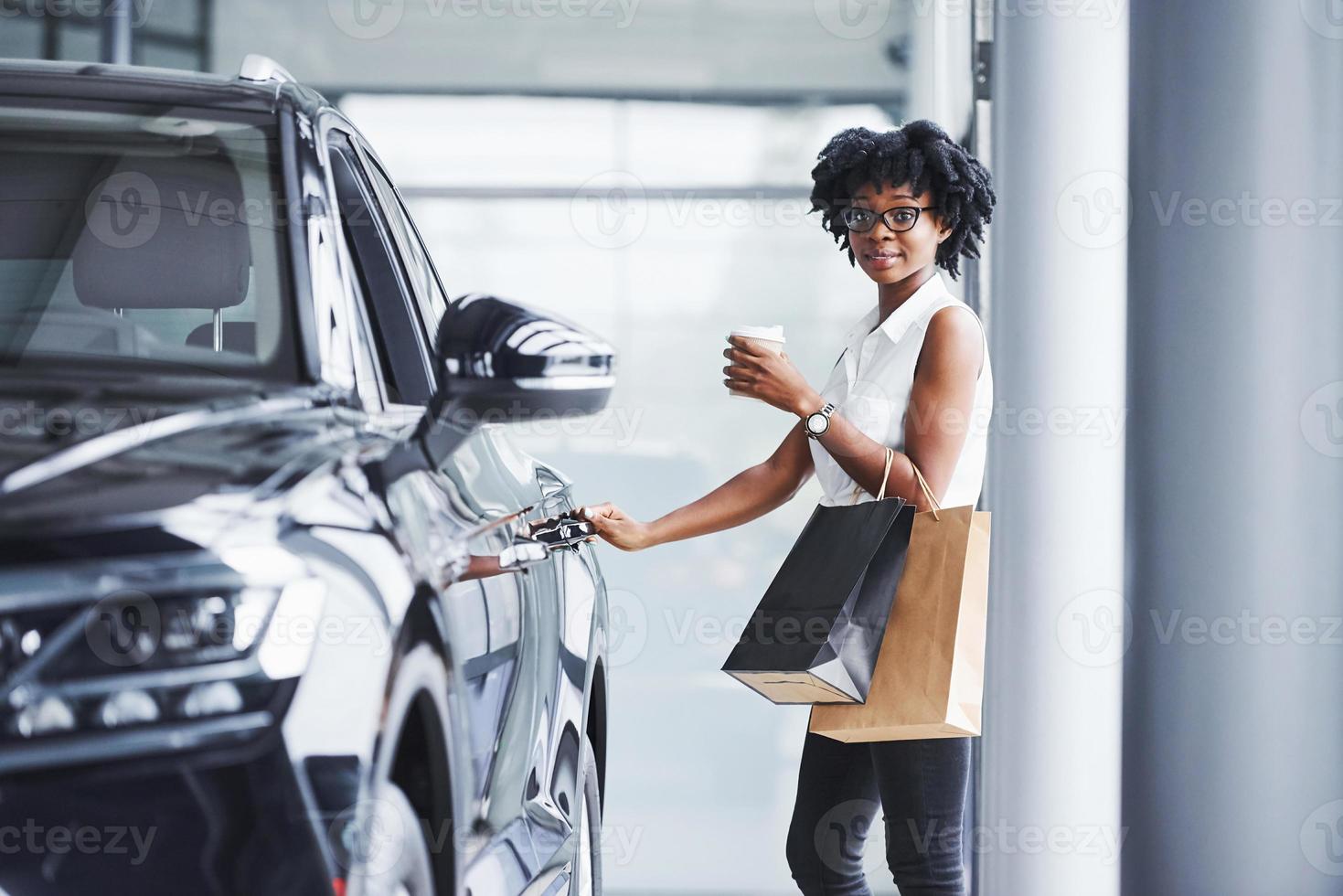 Young african american woman in glasses stands in car salon near vehicle with package in hands photo