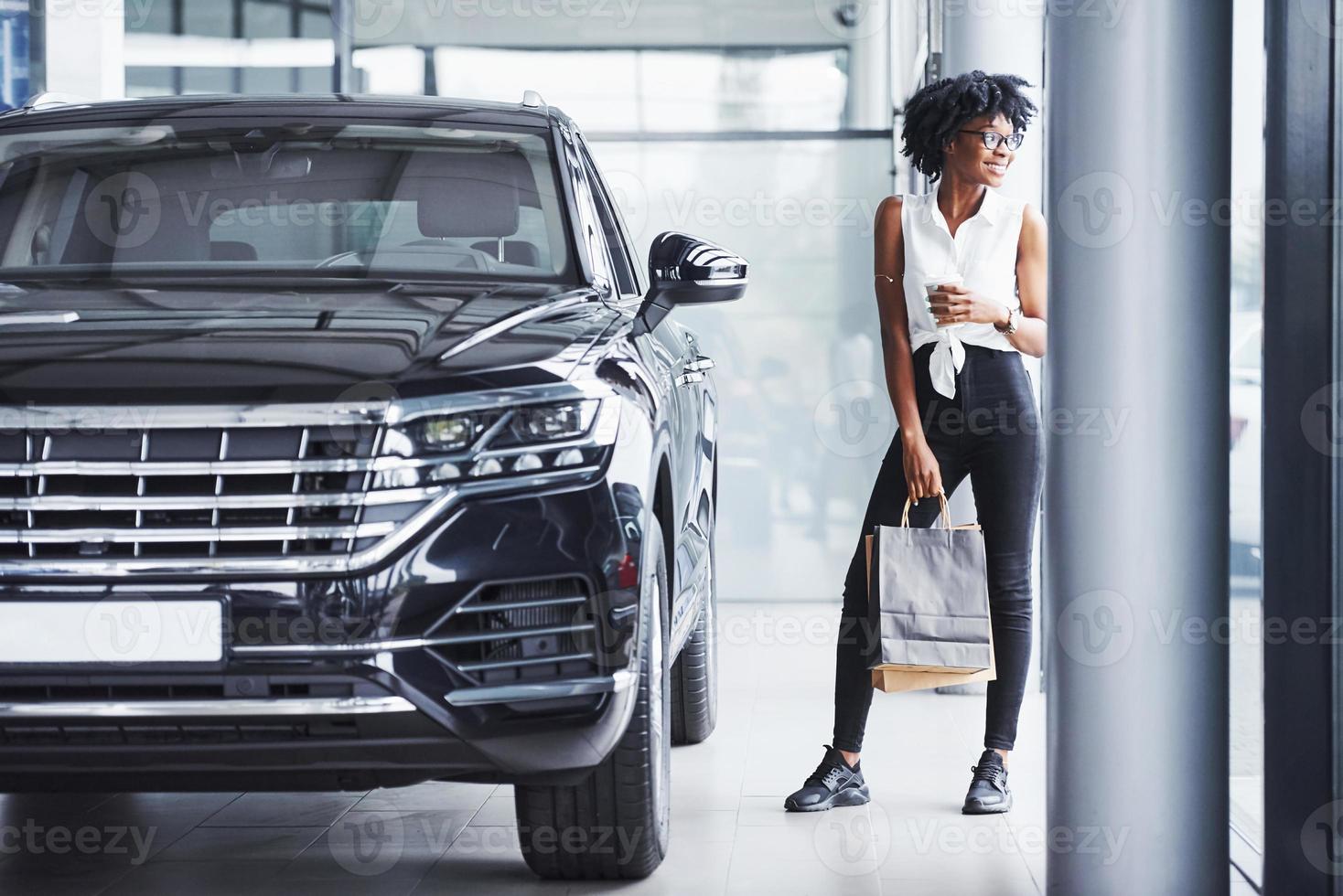 Young african american woman in glasses stands in car salon near vehicle with package in hands photo