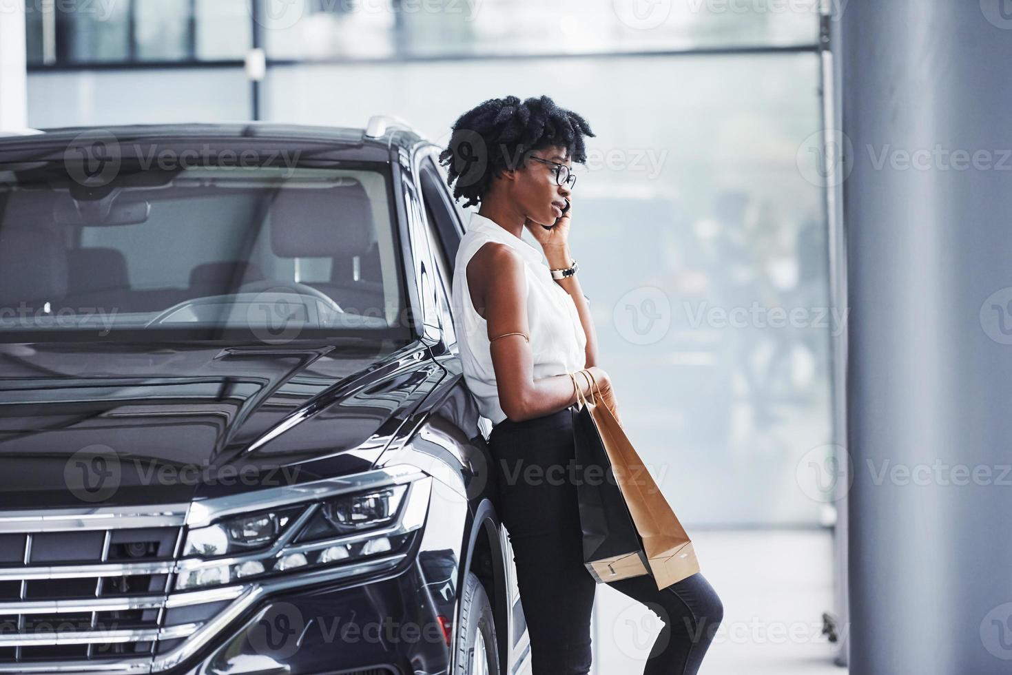 Young african american woman in glasses stands in car salon near vehicle with package in hands photo