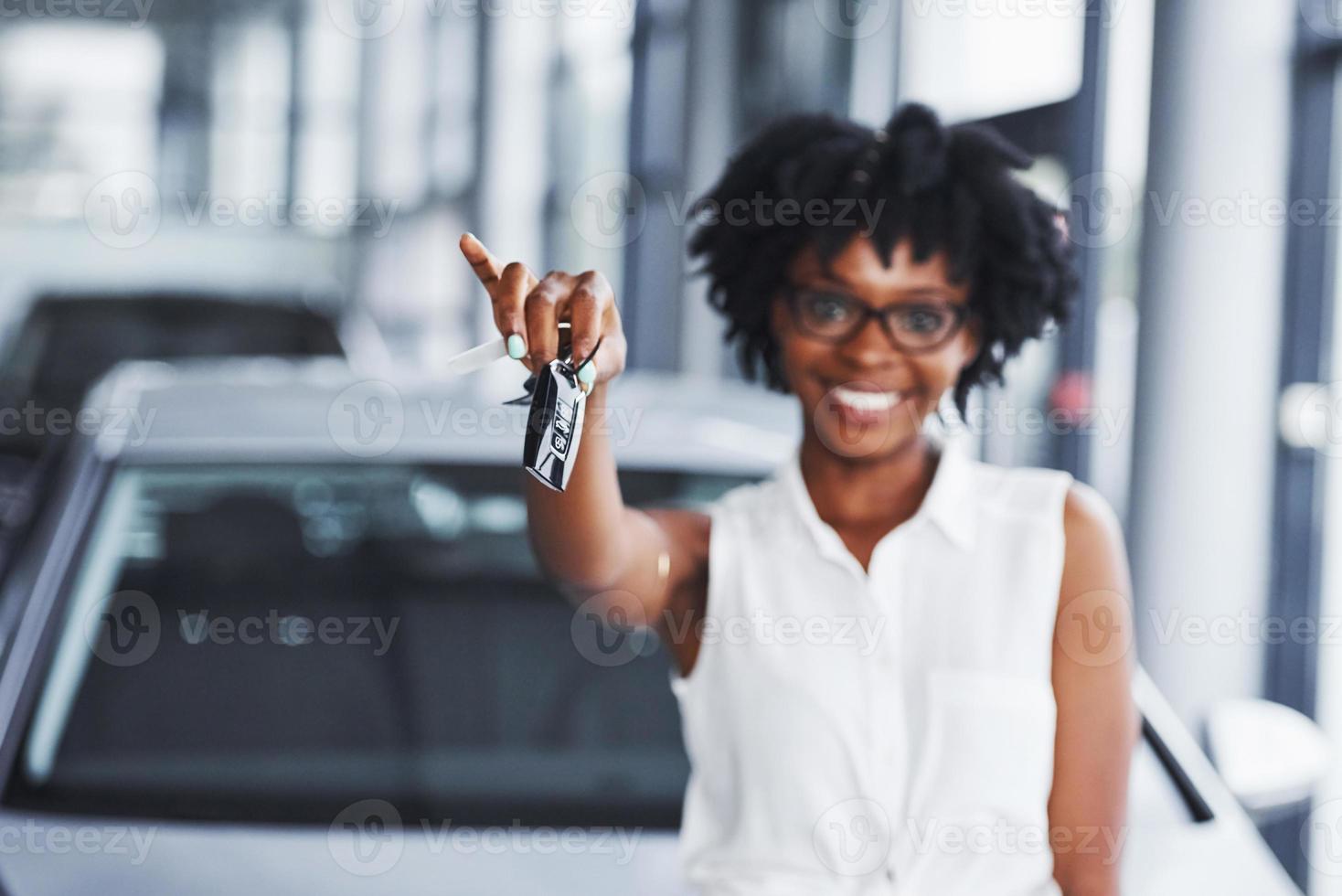 Young african american woman in glasses stands in the car salon near vehicle with keys in hands photo