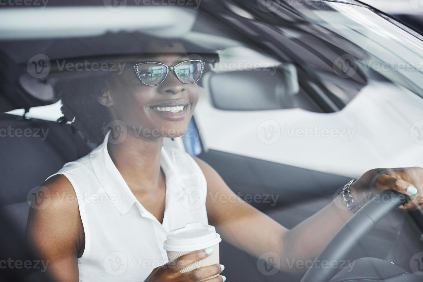 Holds cup of drink. Young african american woman sits inside of new modern car photo