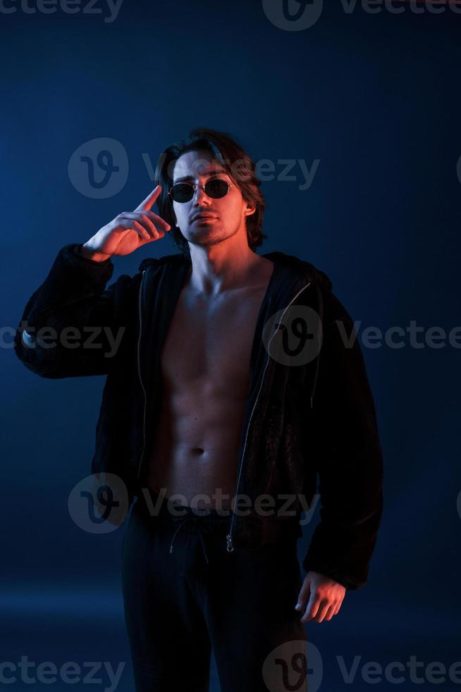 Beautiful man in glasses and black clothes is in the studio with blue neon lighting photo