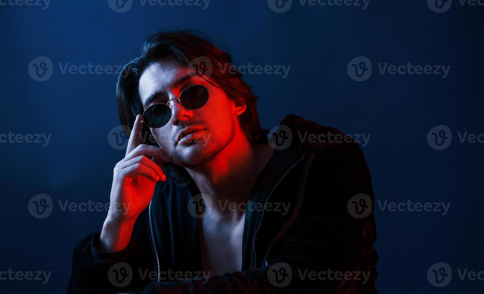 hermoso hombre con gafas y ropa negra está en el estudio con luces de neón azul foto