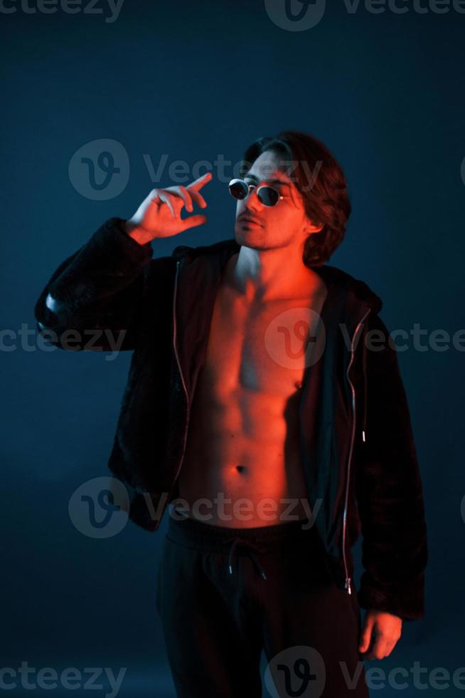 Beautiful man in glasses and black clothes is in the studio with blue neon lighting photo