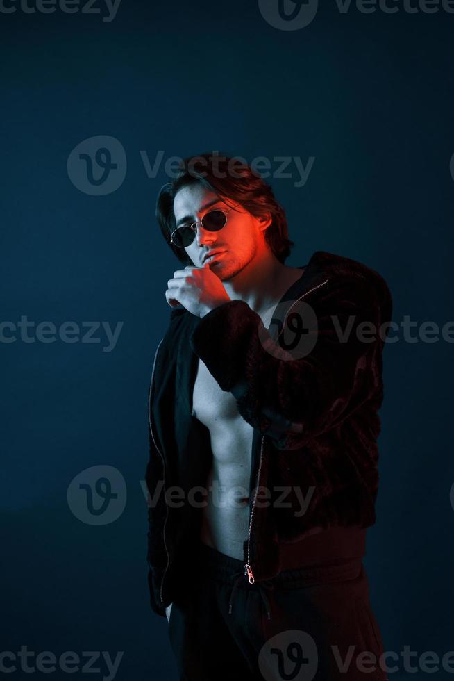 Beautiful man in glasses and black clothes is in the studio with blue neon lighting photo