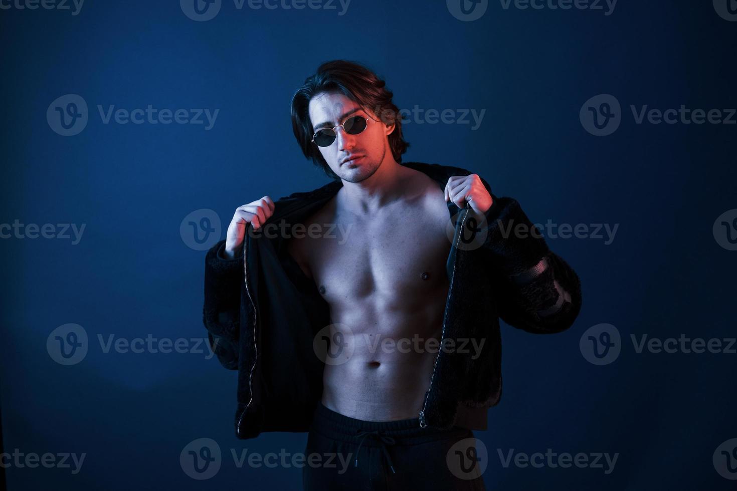 Beautiful man in glasses and black clothes is in the studio with blue neon lighting photo