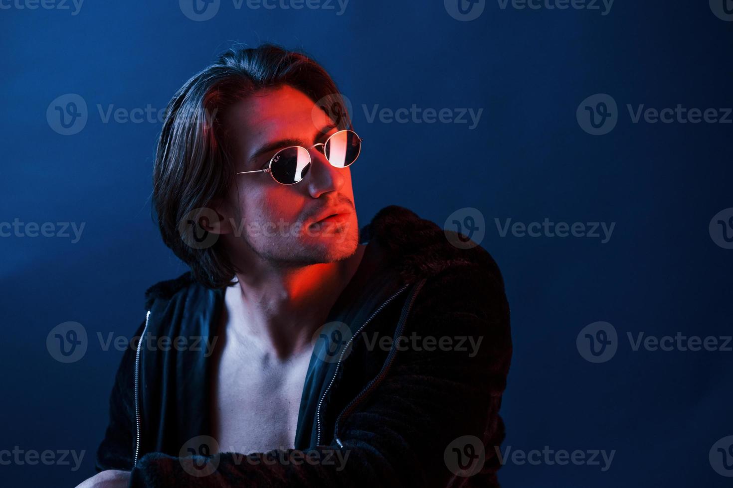 hermoso hombre con gafas y ropa negra está en el estudio con luces de neón azul foto