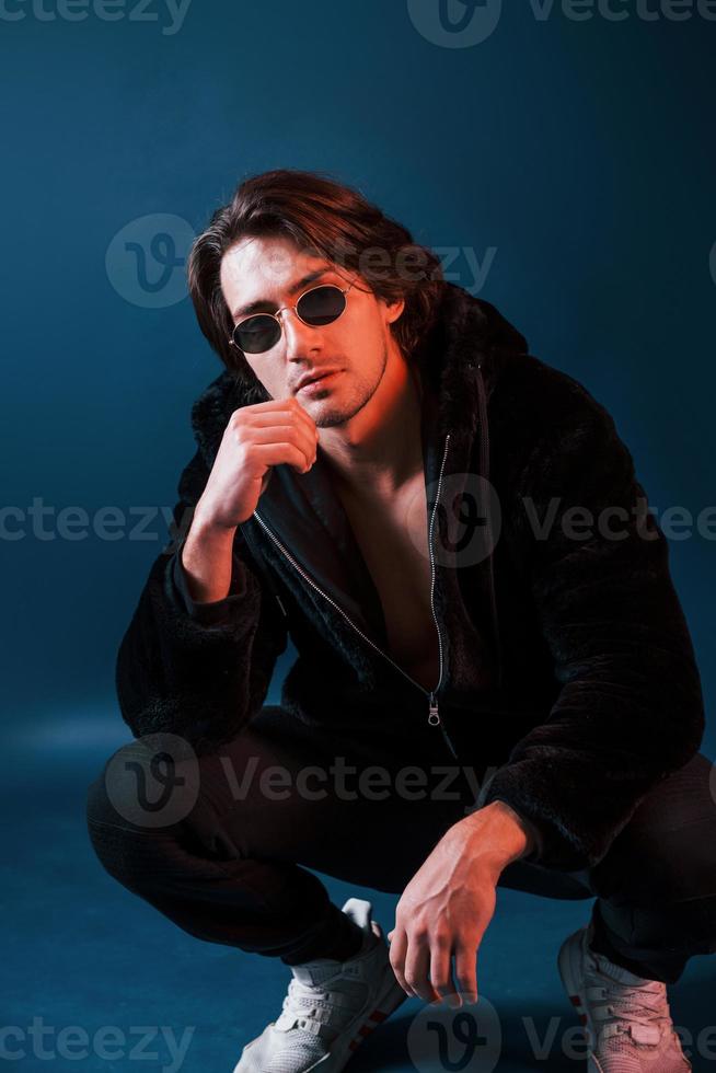 Beautiful man in glasses and black clothes sits in the studio with blue neon lighting photo