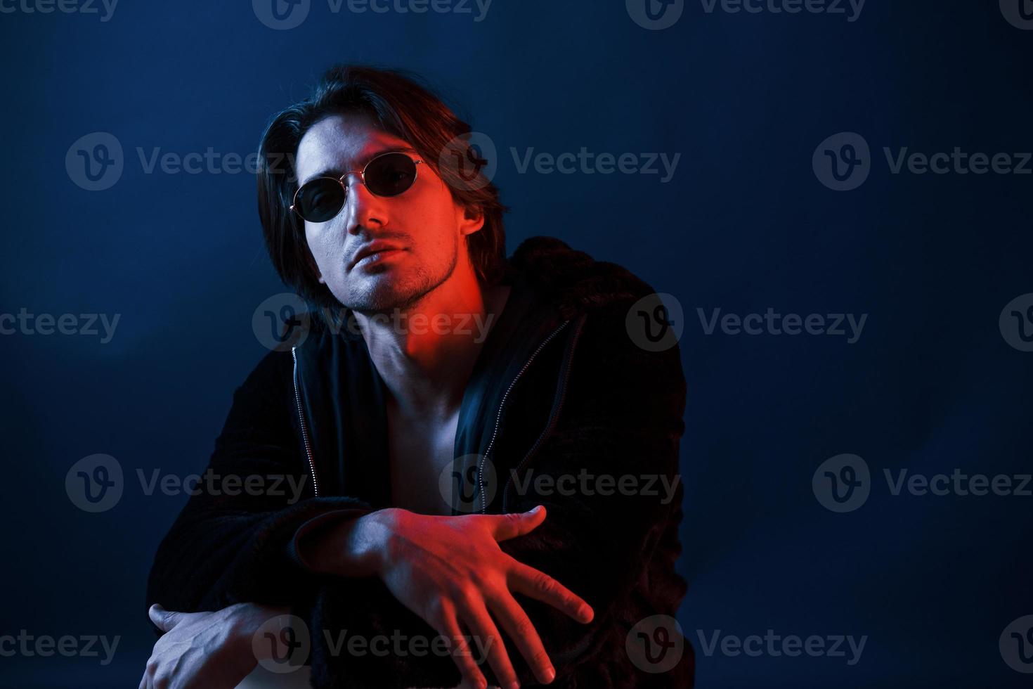 Beautiful man in glasses and black clothes is in the studio with blue neon lighting photo