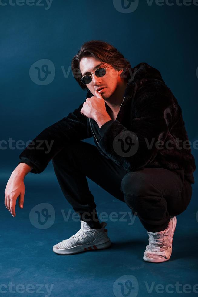 Beautiful man in glasses and black clothes sits in the studio with blue neon lighting photo