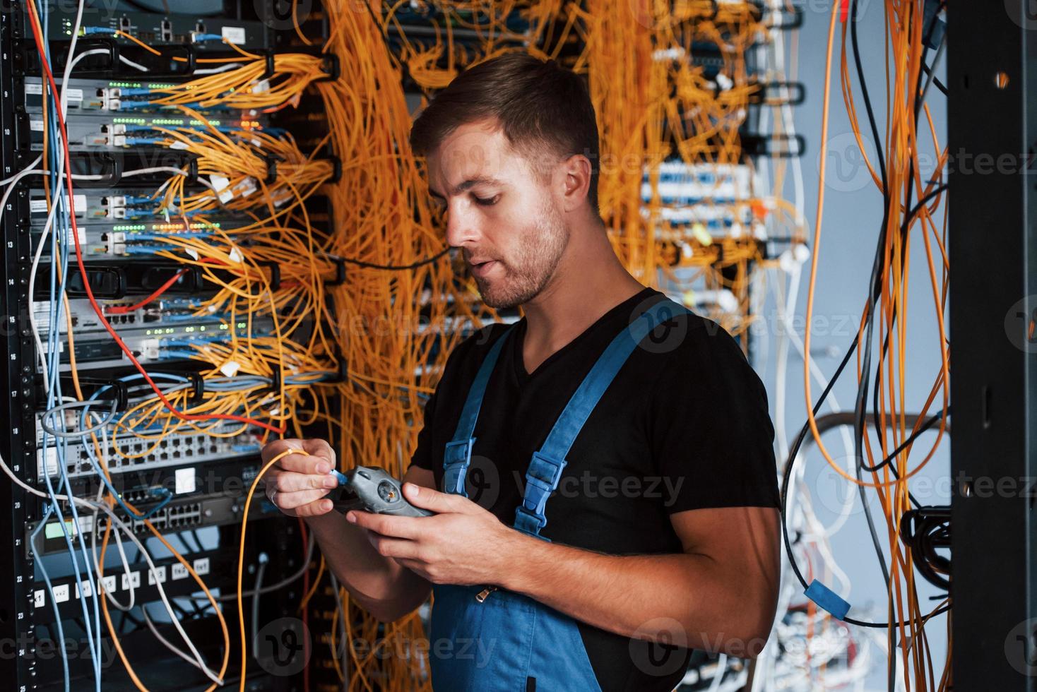 Young man in uniform with measuring device works with internet equipment and wires in server room photo