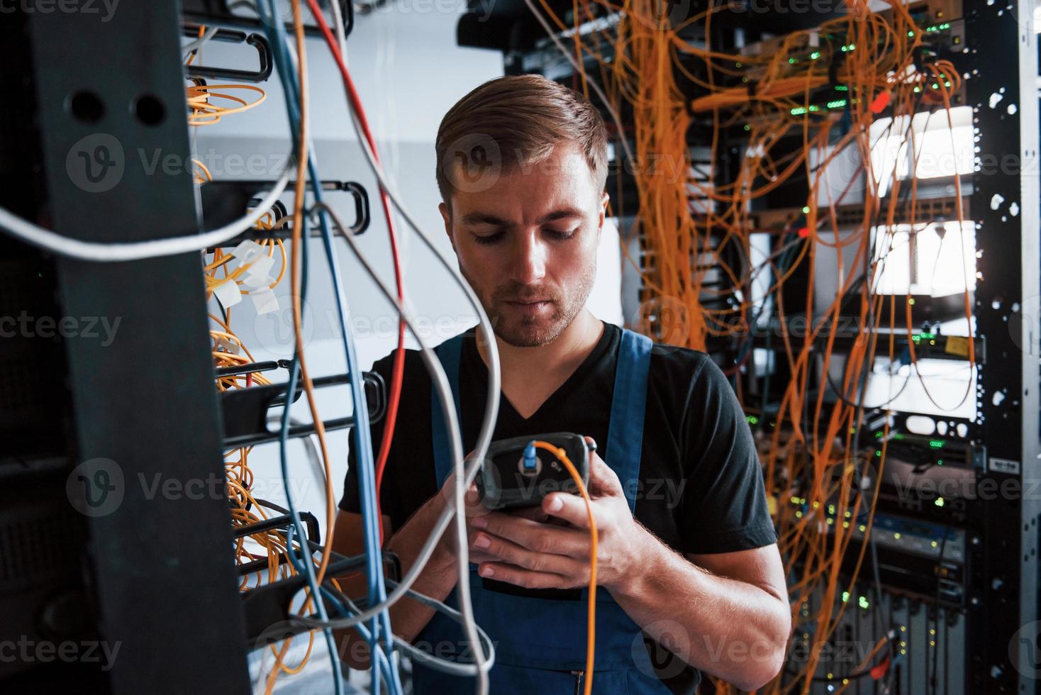 Young man in uniform with measuring device works with internet equipment and wires in server room photo