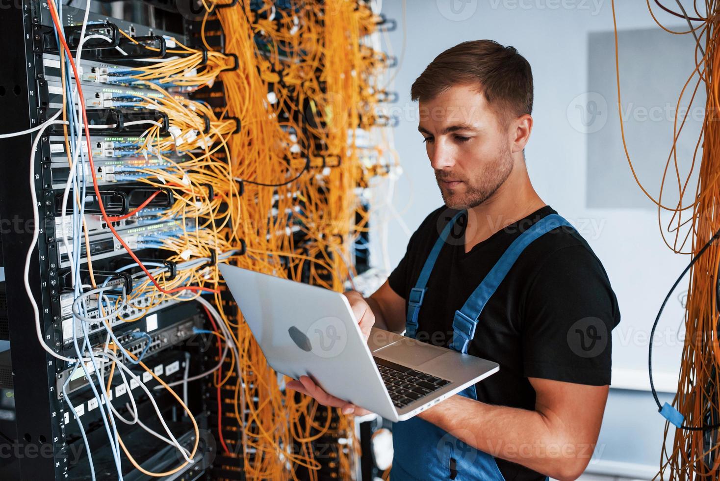 Young man in uniform and with laptop works with internet equipment and wires in server room photo