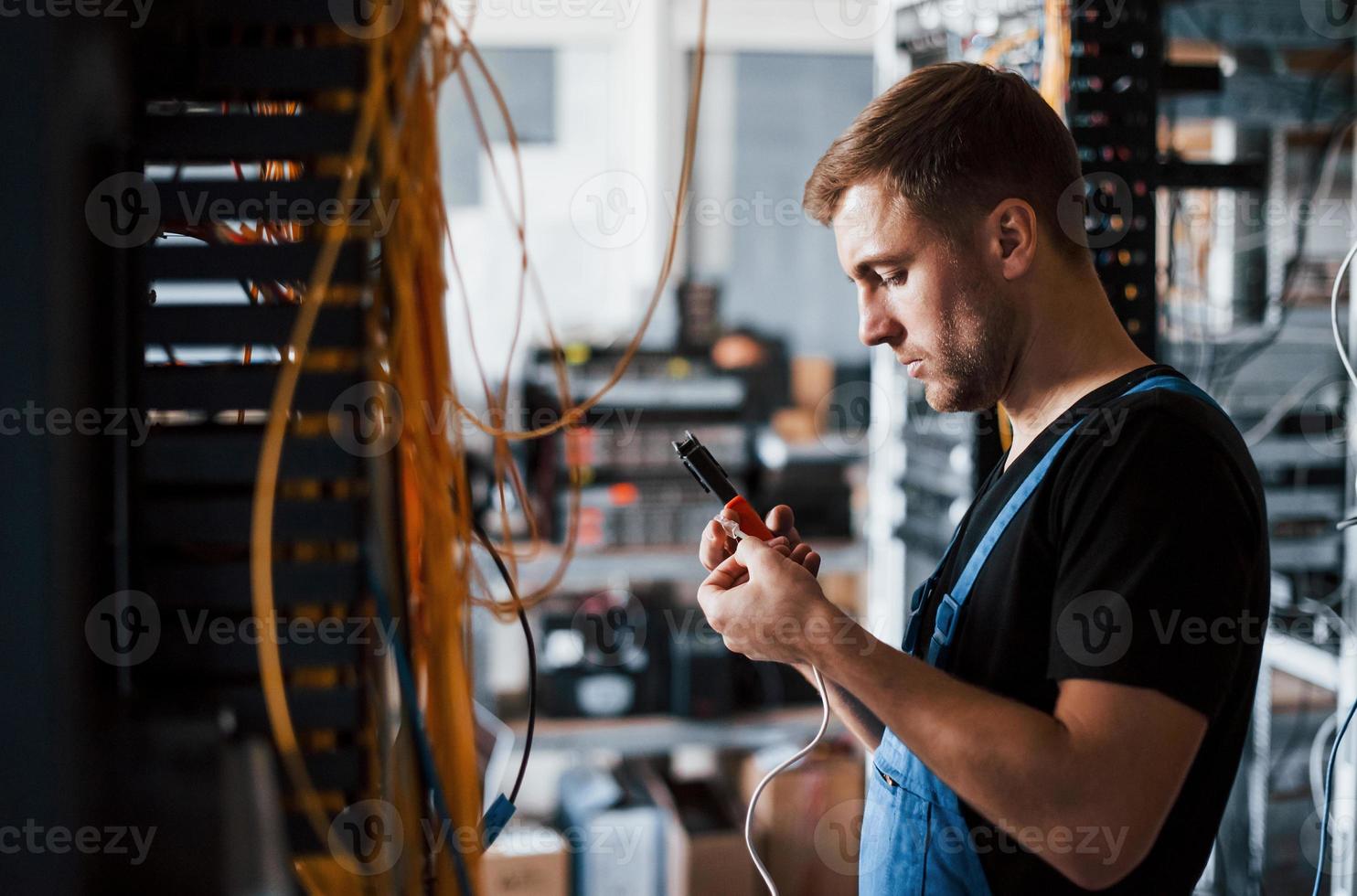 Young man in uniform with measuring device works with internet equipment and wires in server room photo