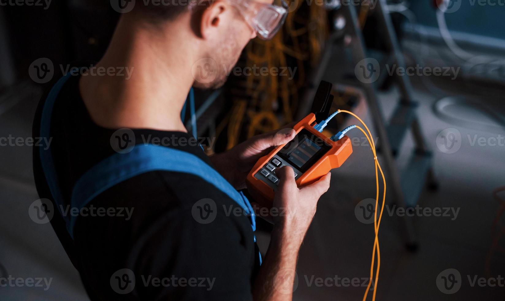Young man in uniform with measuring device works with internet equipment and wires in server room photo