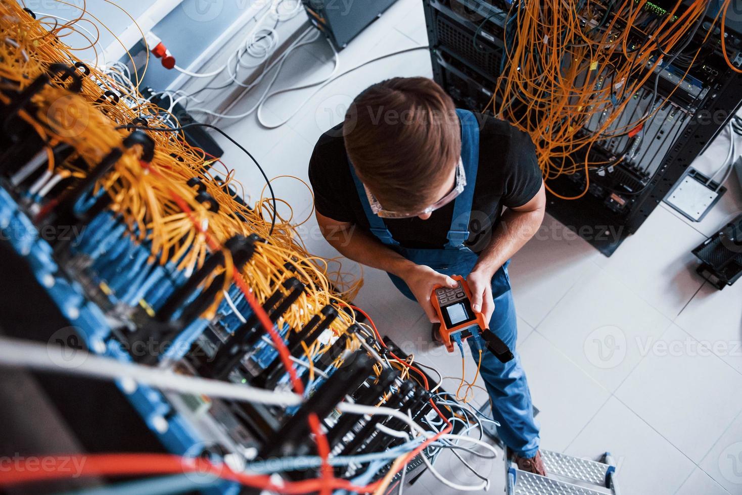 Top view of young man in uniform with measuring device that works with internet equipment and wires in server room photo