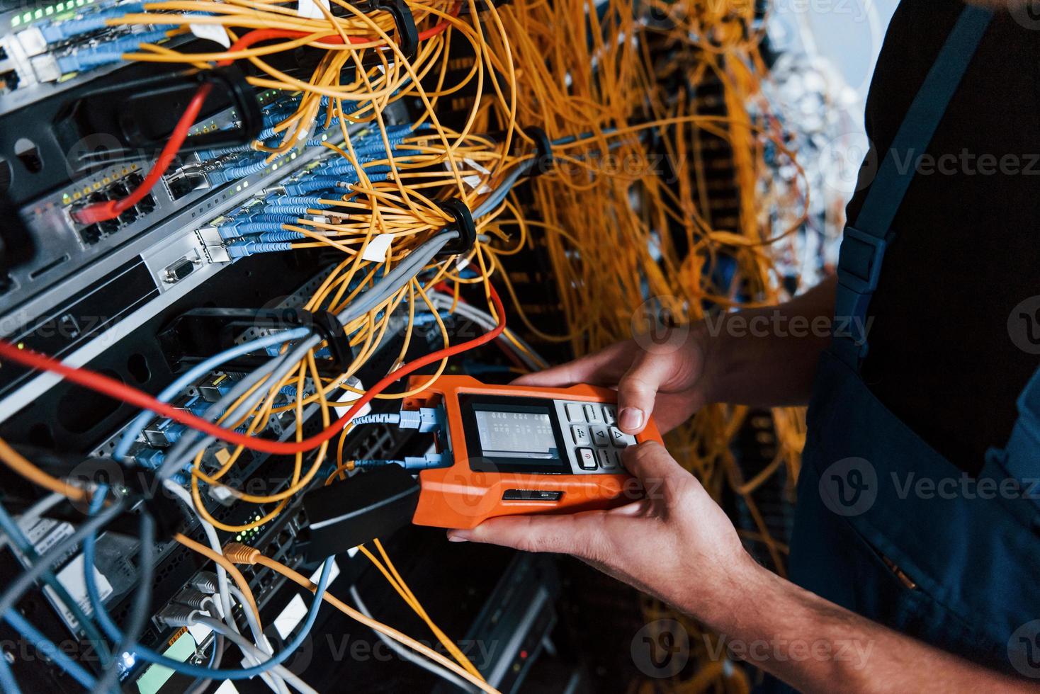 Young man in uniform with measuring device works with internet equipment and wires in server room photo