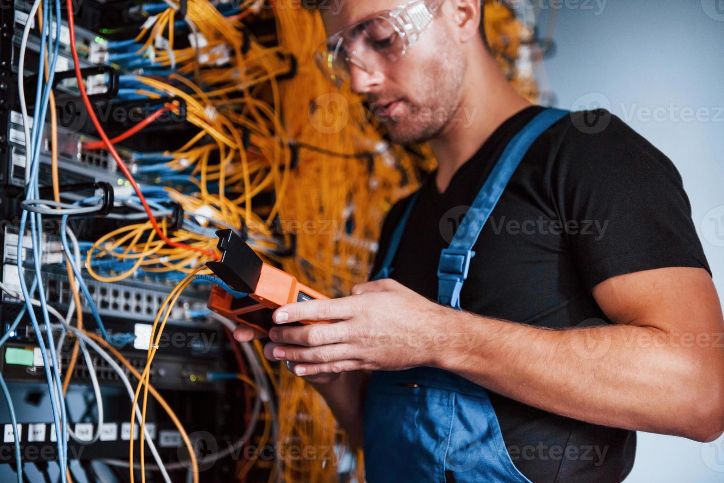 Young man in uniform with measuring device works with internet equipment and wires in server room photo