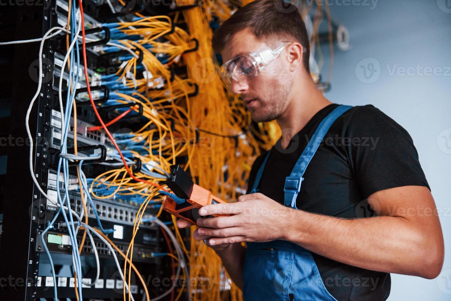 Young man in uniform with measuring device works with internet equipment and wires in server room photo
