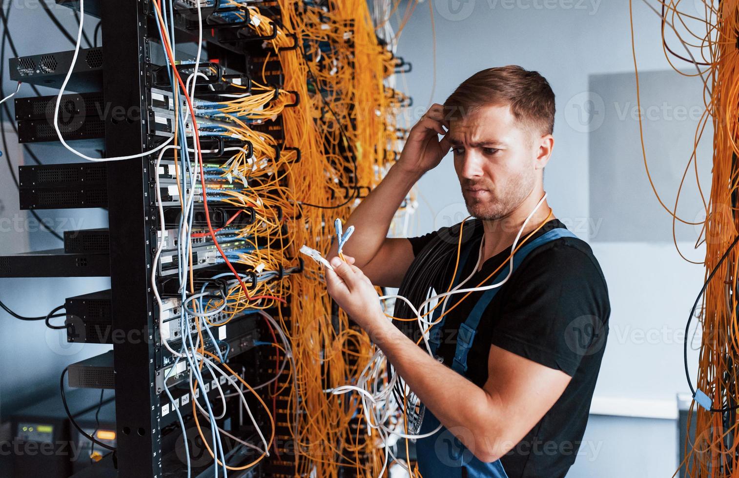 Young man in uniform feels confused and looking for a solution with internet equipment and wires in server room photo