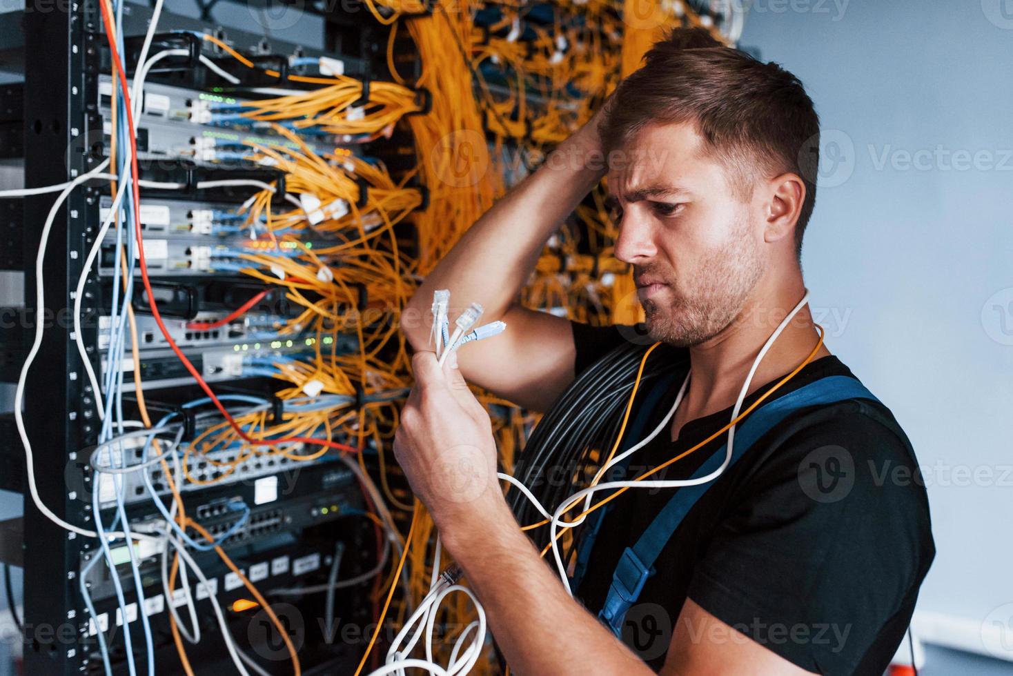 Young man in uniform feels confused and looking for a solution with internet equipment and wires in server room photo