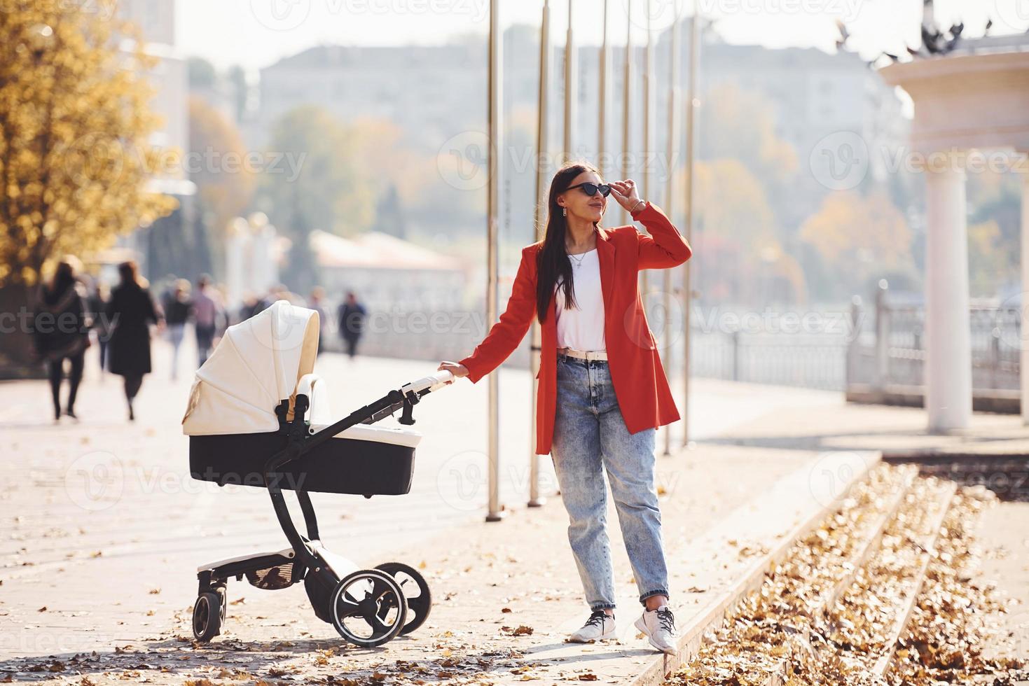 Mother in red coat have a walk with her kid in the pram in the park at autumn time photo
