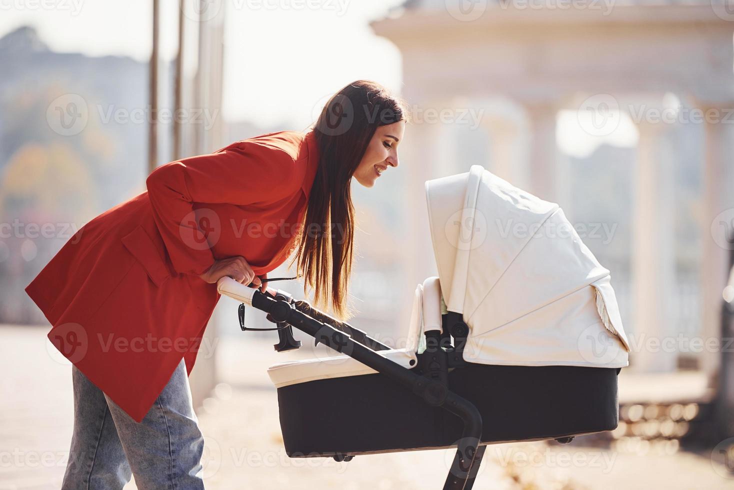 Mother in red coat have a walk with her kid in the pram in the park at autumn time photo