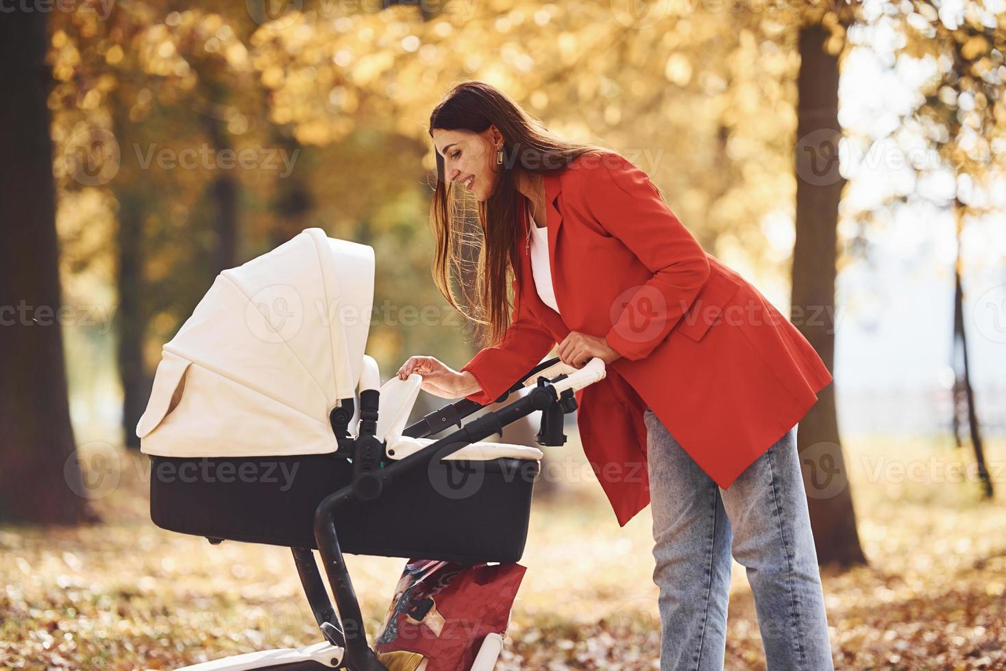 madre con abrigo rojo da un paseo con su hijo en el cochecito en el parque con hermosos árboles en otoño foto