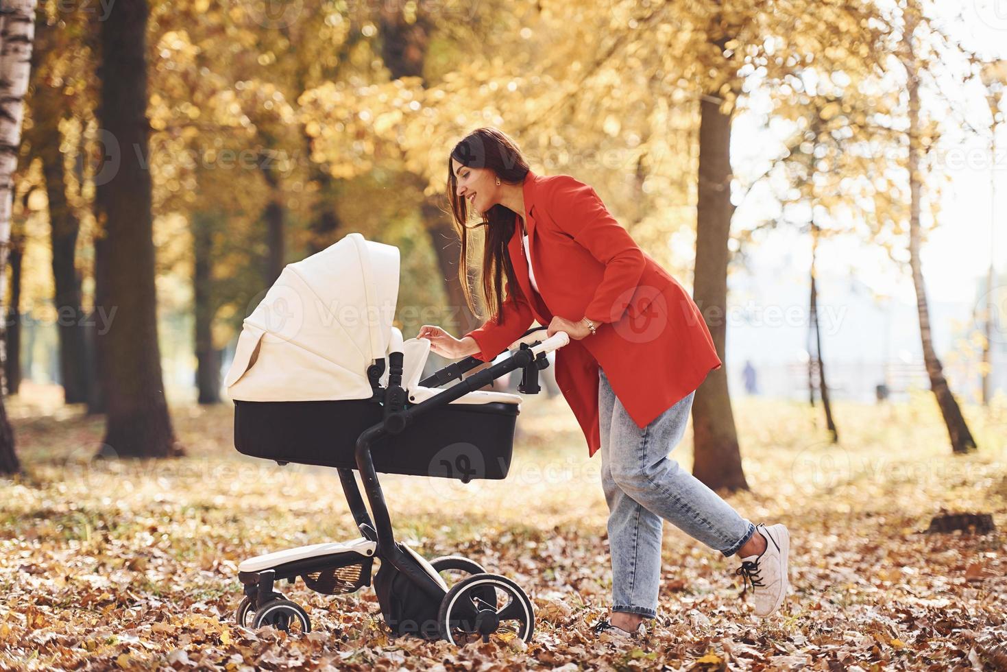 Mother in red coat have a walk with her kid in the pram in the park with beautiful trees at autumn time photo